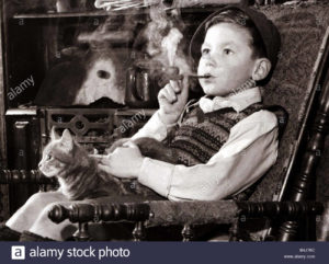 A young boy smoking a pipe and stroking a cat, sitting in a chair looking into the distance.